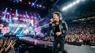 Macklemore performing on a massive concert stadium stage with crowd and lights