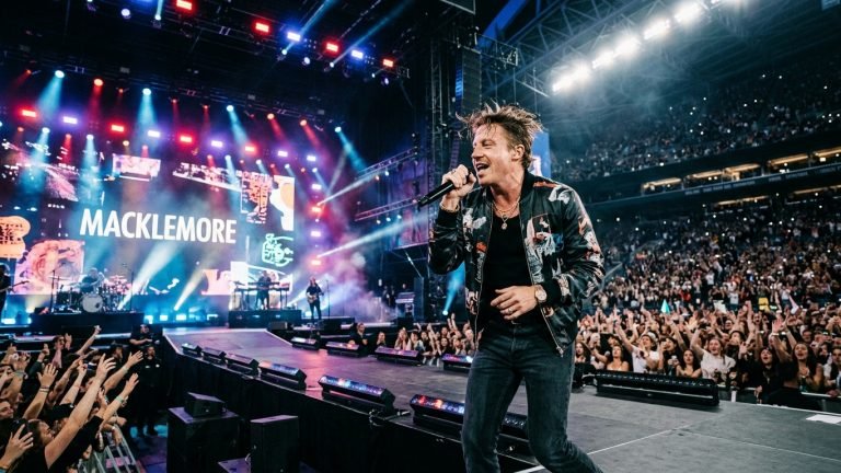 Macklemore performing on a massive concert stadium stage with crowd and lights